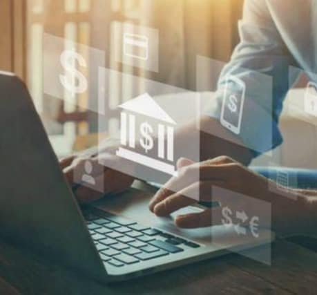 Image of a person typing on a laptop, with overlay graphics of financial icons including a bank symbol, dollar sign, smartphone, and currency symbols, representing digital banking and financial services. The image is illuminated by natural light coming through a window in the background