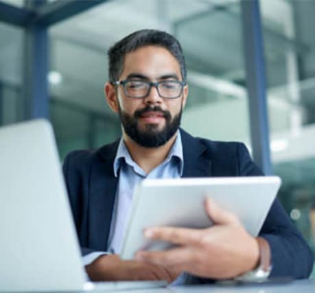 Image of a man with glasses and a beard, sitting at a desk using a tablet. A laptop is open beside him, and the background shows a modern office space with glass walls. The man appears focused, likely engaged in work or a business task