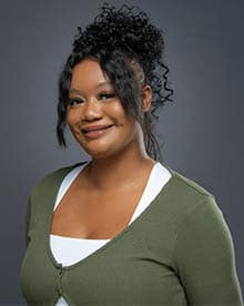 A woman with curly black hair styled in a loose bun smiles gently at the camera. She is wearing a green cardigan over a white top and has a warm, welcoming expression. The background is a plain, neutral gray.