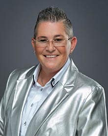 A person with short, styled hair and glasses smiles confidently at the camera. They are wearing a shiny silver blazer over a white button-down shirt. The background is a plain, neutral gray.