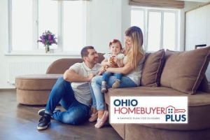 Ohio homebuyers plus a happy family consisting of a man, woman, and a young child sits together on a brown sectional sofa in a bright living room. The man is kneeling on the floor, the woman is sitting on the sofa, and the child is sitting on the woman's lap holding a stuffed animal. There is a flower vase on the window sill in the background. The logo of Ohio Homebuyer Plus is in the bottom right corner of the image.