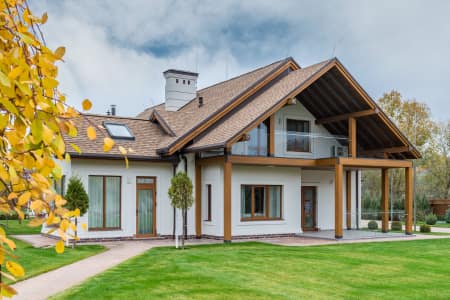 Homepossible modern two-story house with wooden beams and large windows, surrounded by a green lawn and trees under a partly cloudy sky.